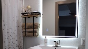 Une salle de bain avec un lavabo blanc, un miroir avec éclairage intégré, un porte-serviettes et un rideau de douche.
