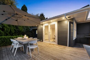 A patio with a table and chairs, an umbrella, and a sliding glass door leading to a kitchen. - Epic Escape - Swim Spa, Redwoods, Sleeps 8 (Rotorua)