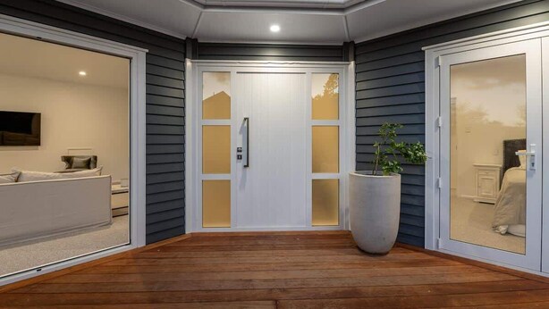 Una casa moderna con una terraza de madera, una puerta blanca con paneles de vidrio y una planta grande en maceta.