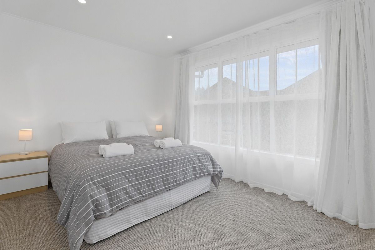 A bedroom with a bed, two bedside tables, a window with sheer curtains, and a carpeted floor.