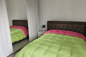 Slaapkamer met twee bedden, elk met een roze en groene dekbedovertrek, een nachtkastje met een lamp en een wit gordijn.
