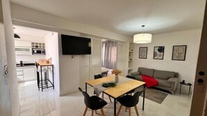 Una sala de estar moderna con un sofá gris, una mesa de comedor de madera con cuatro sillas, un televisor montado en la pared y una cocina visible a través de una puerta abierta.