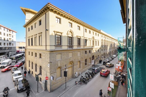 A multi-story building with classical architecture, a street with parked cars and motorcycles, and a clear sky.