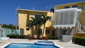 En swimmingpool med en træterrasse foran, omgivet af en gul bygning med en trappe og et lille haveområde.