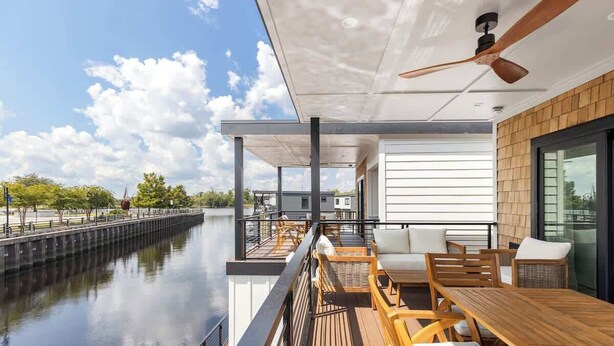 Una terraza moderna al aire libre con mobiliario de madera, un ventilador de techo y vistas a un cuerpo de agua tranquilo.