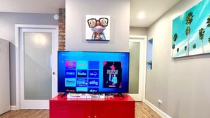 Un saló amb televisió de pantalla plana sobre un suport vermell, un quadre penjat a la paret amb un personatge de dibuix animat, i una pintura de palmeres.