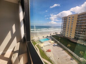 Vista desde una ventana sobre una playa, una piscina y un estacionamiento con un coche rojo.