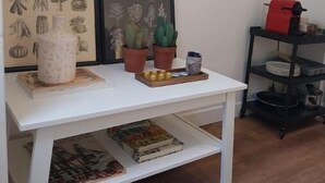 Ein weißer Tisch mit einem gerahmten Pflanzenprint, einer Topfpflanze und einer Ablage mit Gegenständen. Rechts daneben befindet sich ein rotes Objekt auf einem schwarzen Regal.