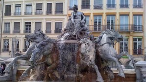 Ein klassischer Brunnen mit einer sitzenden Figur auf der Spitze und Pferden an jeder Seite, vor einer Fassade mit zahlreichen Fenstern und Balkonen.