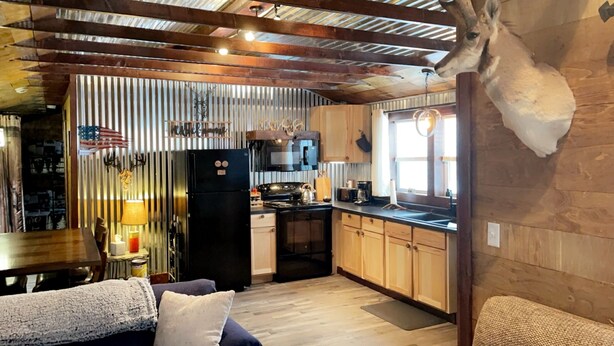 Una cocina compacta con armarios de madera, un refrigerador negro y un microondas. Hay una mesa de comedor con sillas y una lámpara.