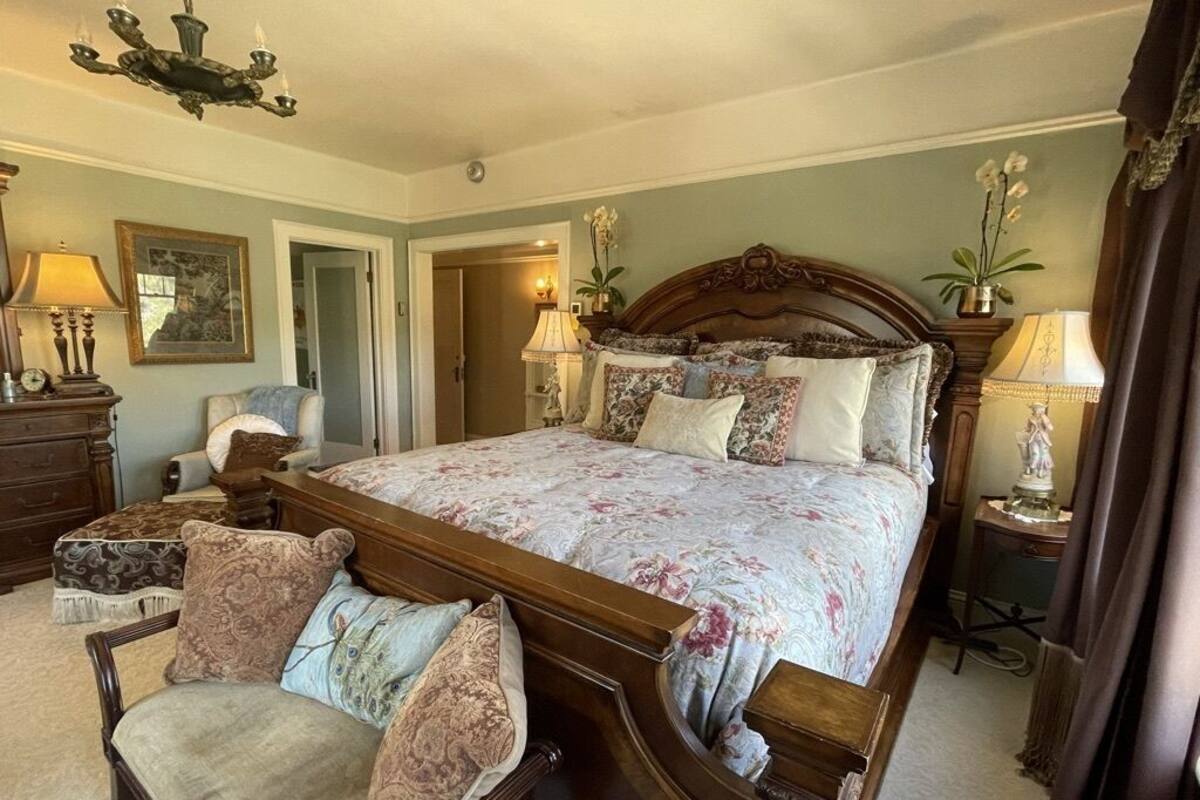 A bedroom with a large bed, a wooden nightstand, a chair, and a chandelier.