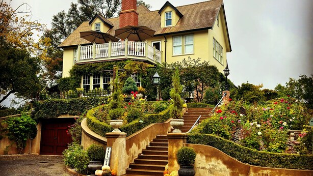 Une grande maison avec un escalier imposant menant à l’entrée, entourée de jardins luxuriants et d’une pelouse bien entretenue.