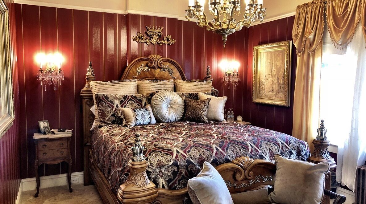 A luxurious bedroom with a large ornate bed, two wall sconces, a chandelier, and a framed picture on the wall.