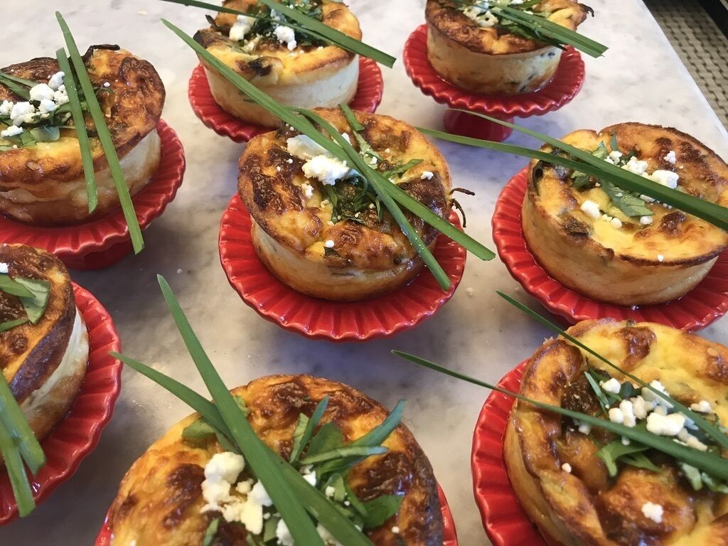 Eine Auswahl an Mini-Quiches in roten Bechern, garniert mit grünen Schnittlauchhalmen und zerbröseltem Käse.