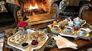 Un salon chaleureux avec une cheminée, une table à manger dressée avec du thé et des pâtisseries, et un canapé agrémenté de coussins décoratifs.