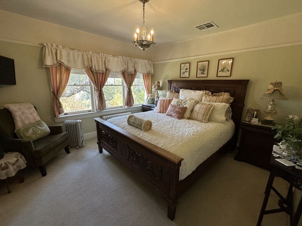 A bedroom with a large bed, a wooden headboard, a chandelier, a window with curtains, a radiator, a bedside table, and a chair.