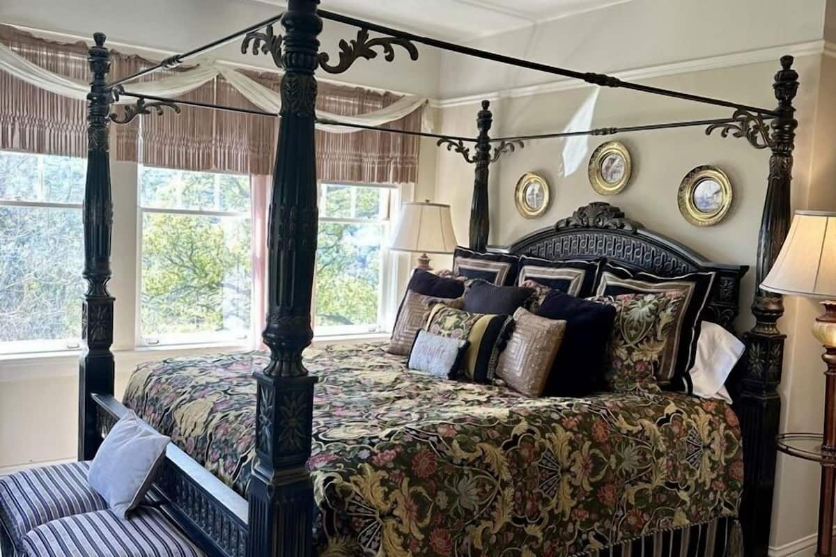 A four-poster bed with a floral bedspread, a bench, and a nightstand with a lamp.