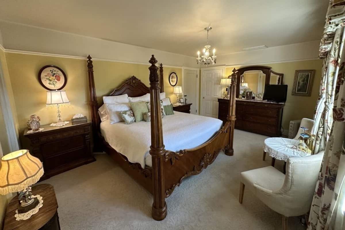 A bedroom with a four-poster bed, a chandelier, a dresser, a chair, and a small table with a lamp.