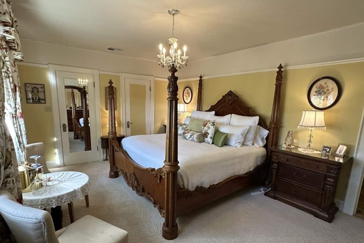 A four-poster bed with a chandelier, a bedside table with a lamp, and a chair with a table.