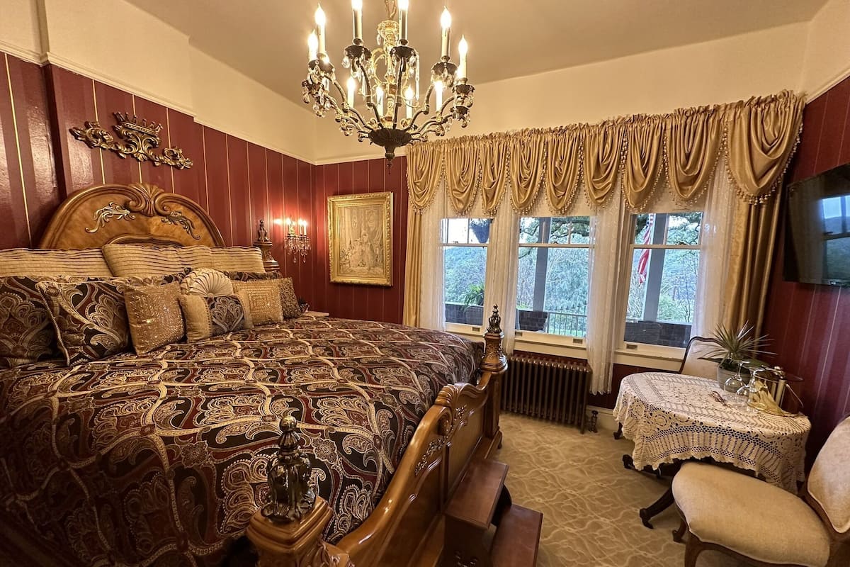 A traditional bedroom with a large bed, ornate wooden headboard, a chandelier, a small table with a lace tablecloth, and a chair.