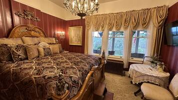Un dormitorio tradicional con una cama grande, un cabecero de madera ornamentado, una araña, una mesita con mantel de encaje y una silla.