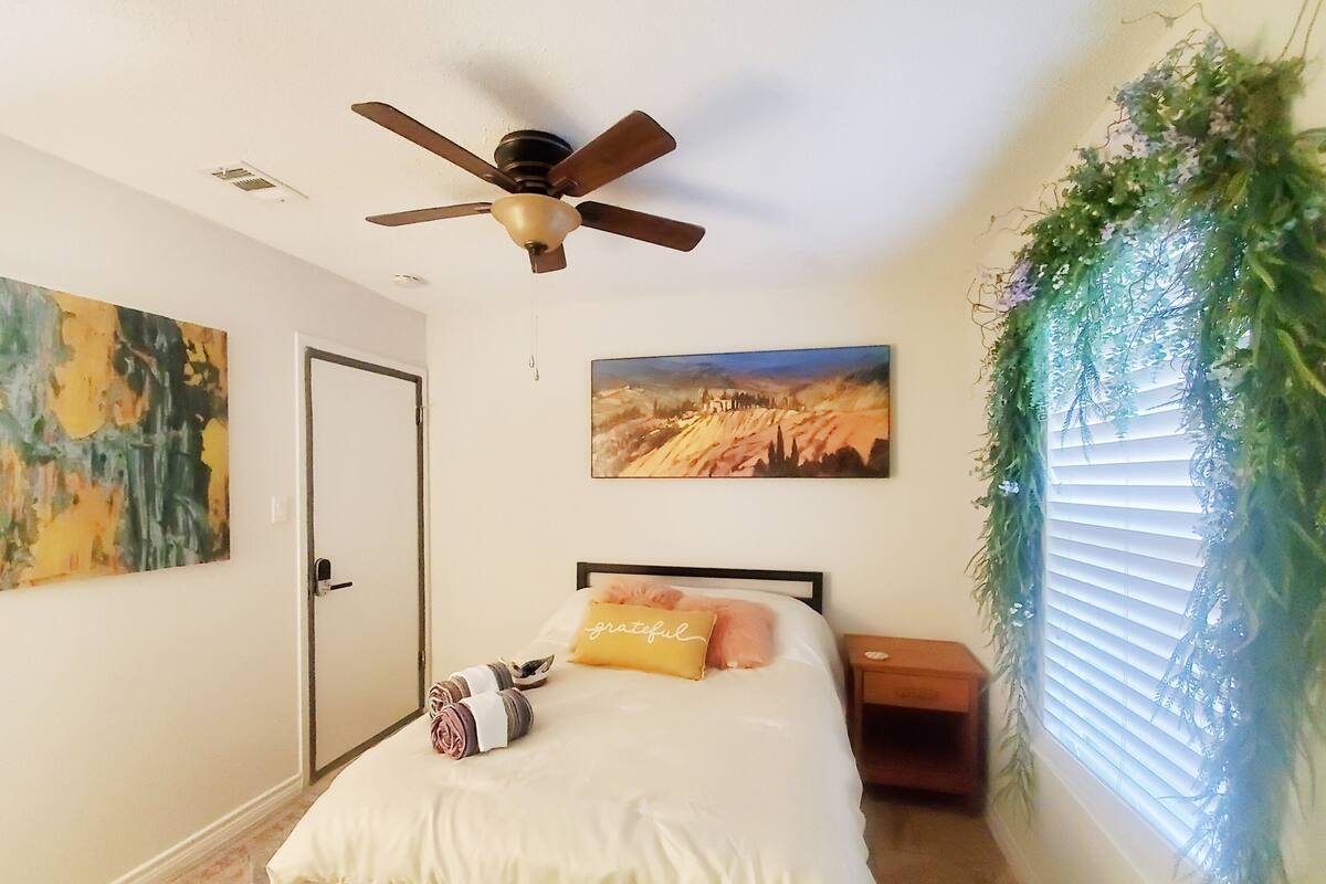A bedroom with a bed, a ceiling fan, a door, and two paintings on the wall.