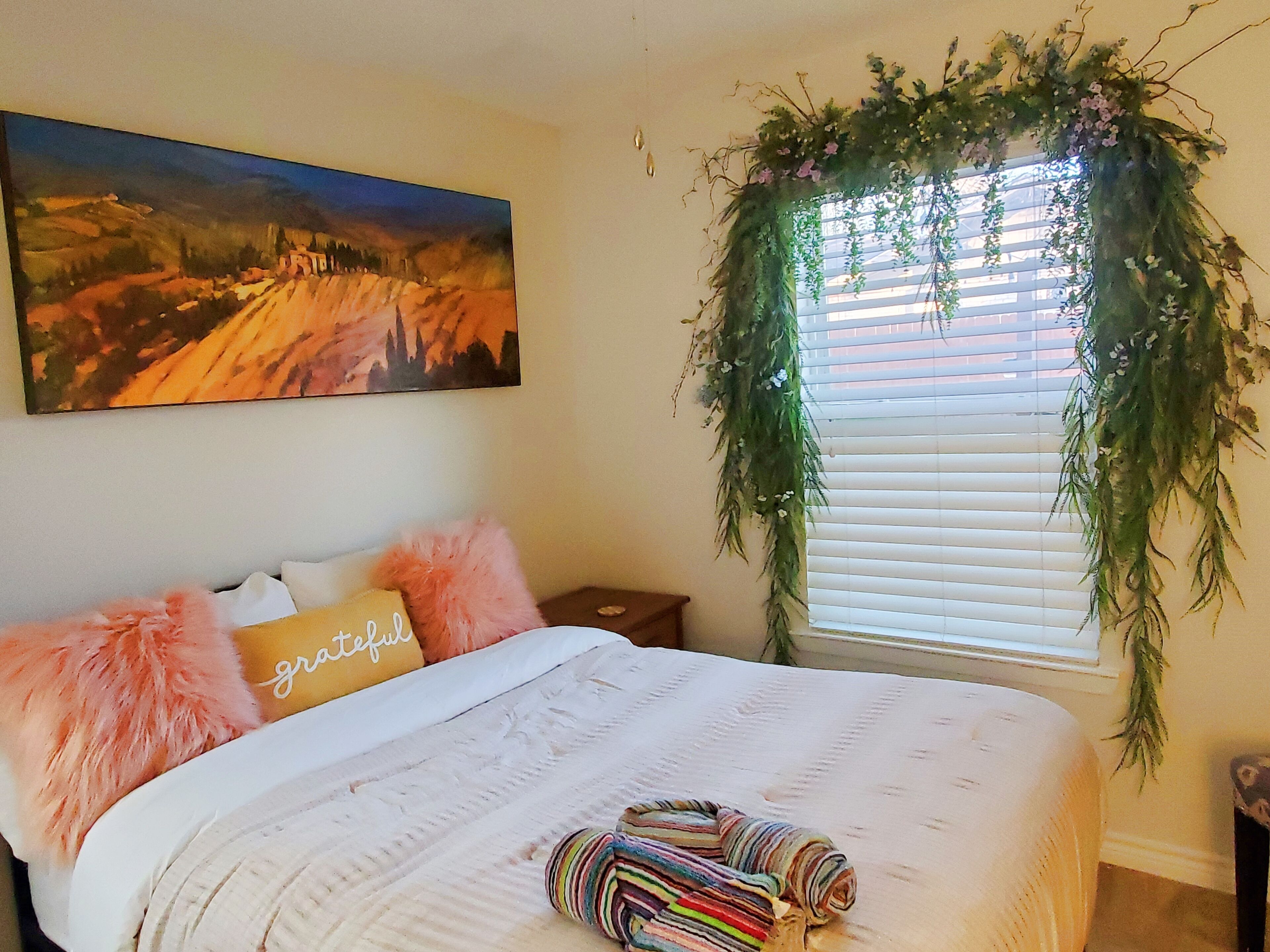 Un dormitorio con cama, almohadas, una manta, un cuadro y una ventana con una planta.