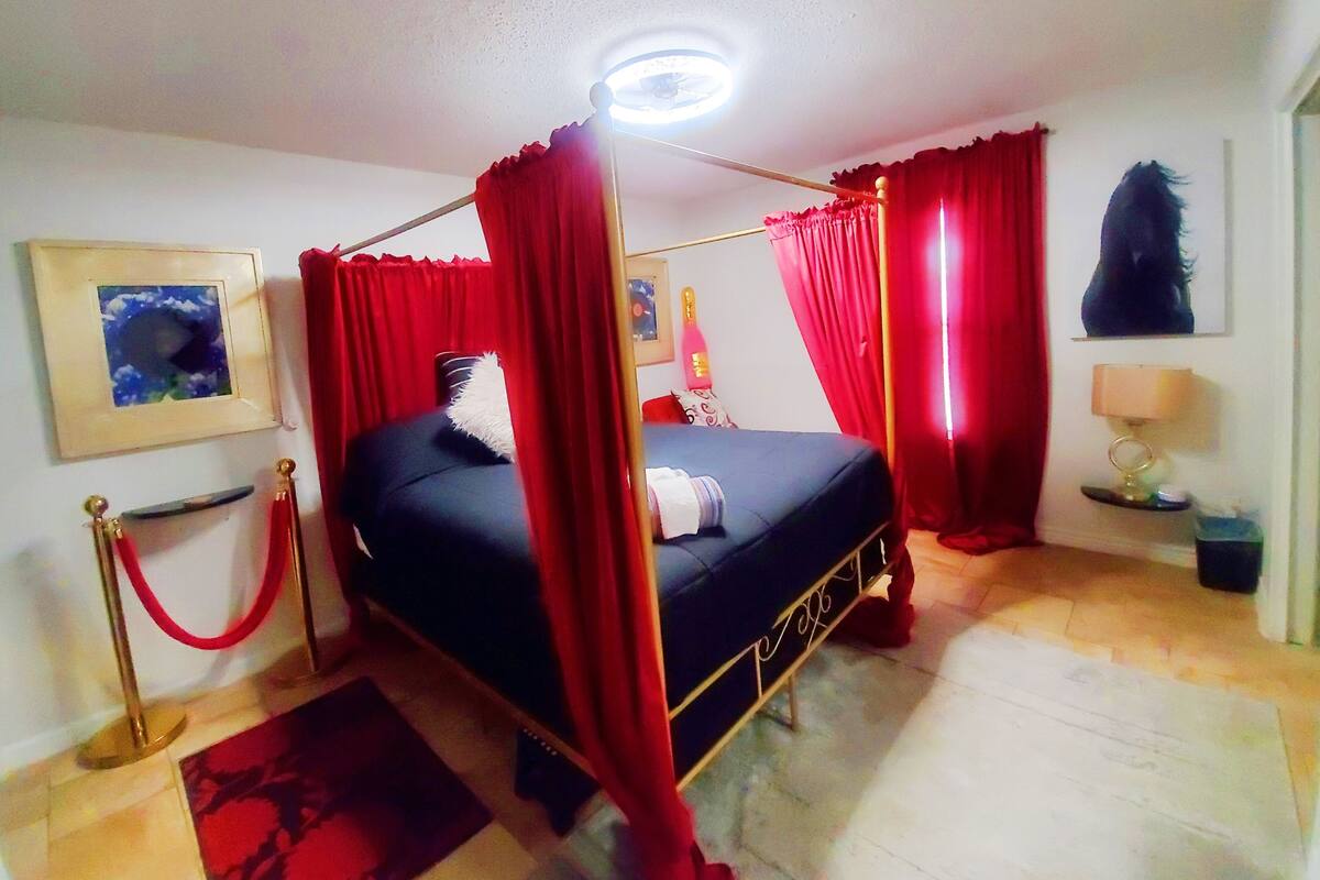 A four-poster bed with red curtains, a bedside table with a lamp, and a framed picture on the wall.