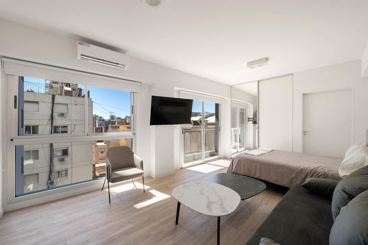 A modern, well-lit room with a sofa, a small table, a TV mounted on the wall, and large windows offering a view of the cityscape.