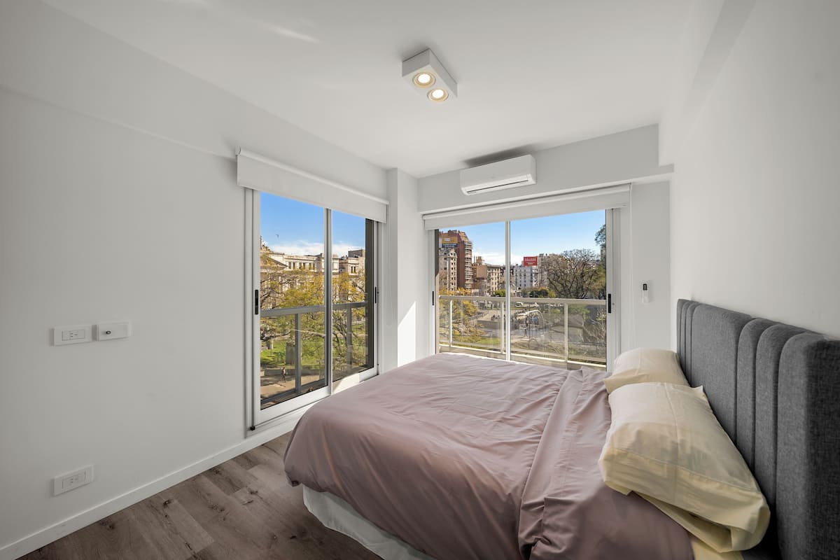 A bedroom with a large bed, a view of the city through floor-to-ceiling windows, and a modern ceiling design.