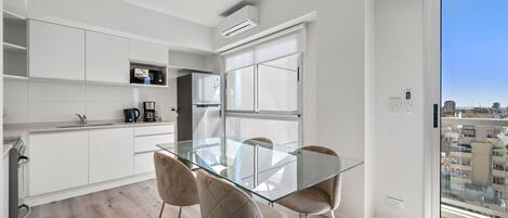 A modern kitchen with white cabinets, a glass-top dining table, and a city view through a large window.
