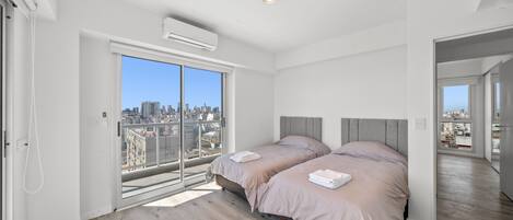 Une chambre moderne avec deux lits, une grande porte-fenêtre coulissante donnant sur un balcon et une vue sur la ville.