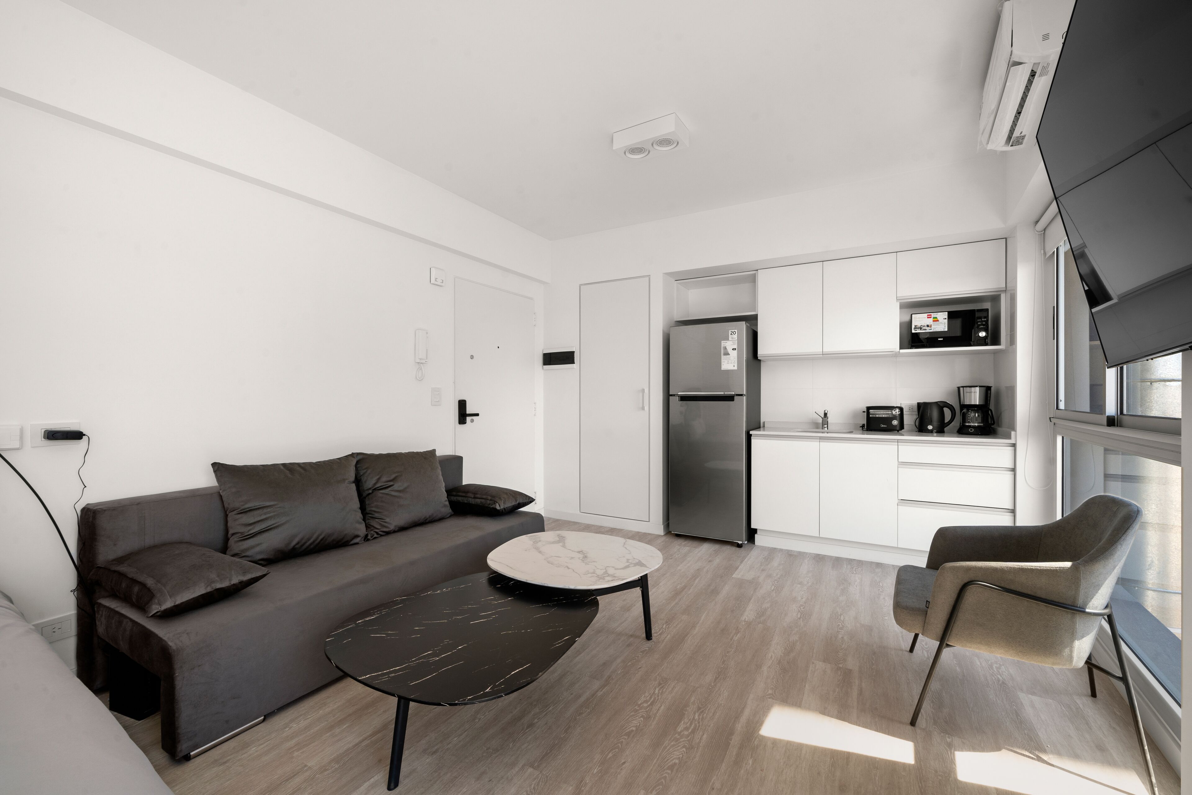 Una sala de estar moderna con un sofá, una mesa de centro y un sillón. Hay una zona de cocina con un refrigerador, microondas y armarios.