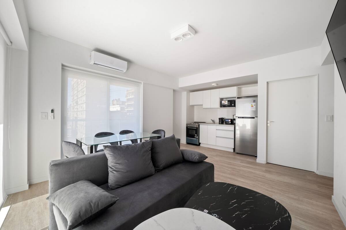 A modern apartment with a grey sofa, a dining table, and a kitchen area.