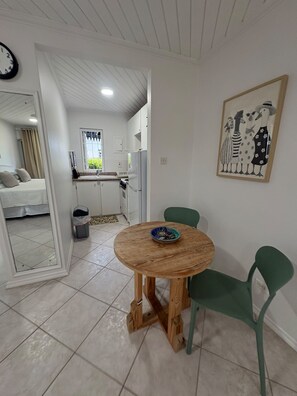 A room with a wooden table, two chairs, a bed, a kitchen area, and a painting on the wall.