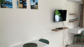 Una habitación con una mesa blanca y dos sillas verdes, un televisor de pantalla plana montado en la pared y un estante con una planta.