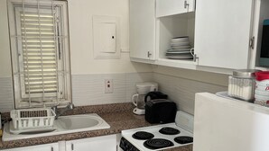 Cocina compacta con armarios blancos, estufa, pileta y ventana con persianas.
