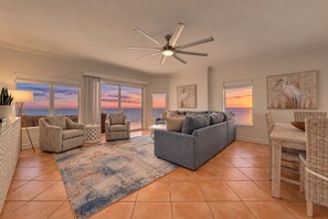 A spacious living room with a large gray sofa, a small armchair, and a dining area with wicker chairs. - Admirals Quarters 1509 | Beachfront Paradise | Penthouse Gulf Views | Beach Getaways (Orange Beach)
