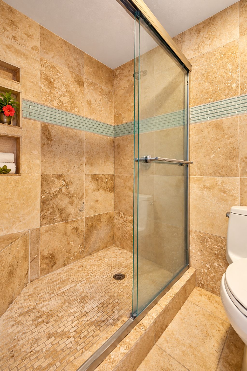 Ein Badezimmer mit einer gläsernen Duschkabine, einer Toilette und einem Regal mit Handtüchern und Blumen.