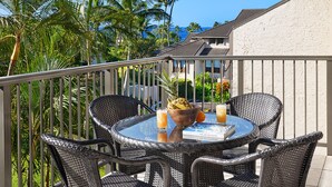 A round glass table with wicker chairs on a balcony, overlooking a tropical landscape with palm trees and a building in the background. - Kahala 433 | Ocean View 1BR at Poipu Kai, Walk to Beach (Koloa)