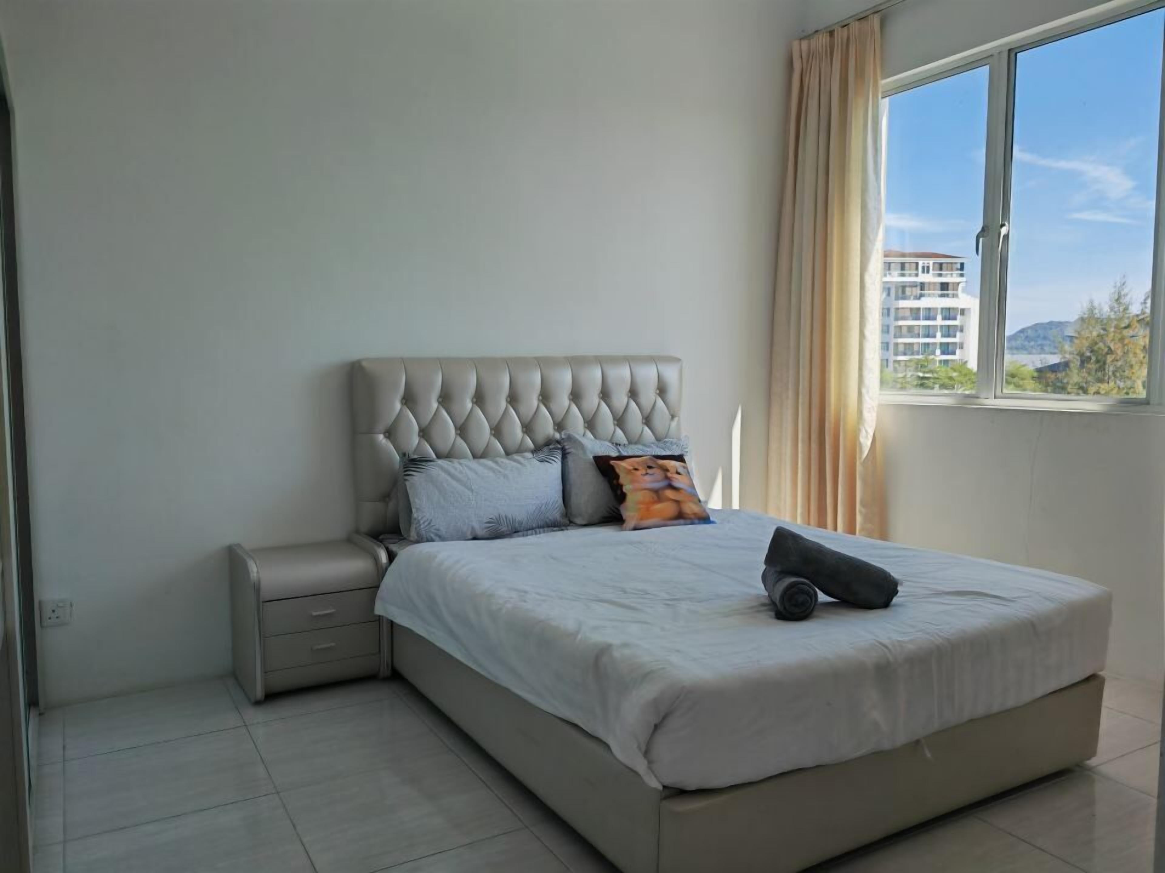 A bedroom with a tufted headboard, a bed with a white bedspread, a nightstand, and a window with a view of buildings and hills.