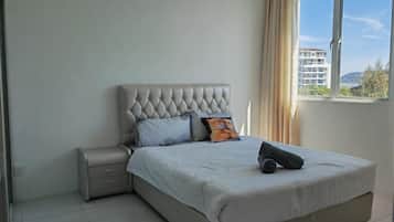 A bedroom with a tufted headboard, a bed with a white bedspread, a nightstand, and a window with a view of buildings and hills.