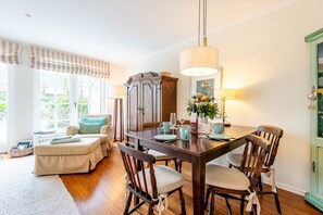 Een eetruimte met een houten tafel, stoelen en een kroonluchter. Achterin staat een bank en een kast.