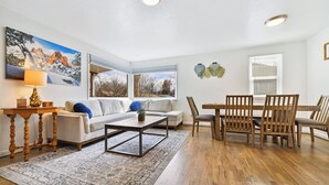 A living room with a sofa, coffee table, and dining table with chairs. There are windows with blinds and a painting on the wall. - ᨒ Hot Tub / Fire Pit / Game Lounge / Fenced Yard ᨒ (Colorado Springs)