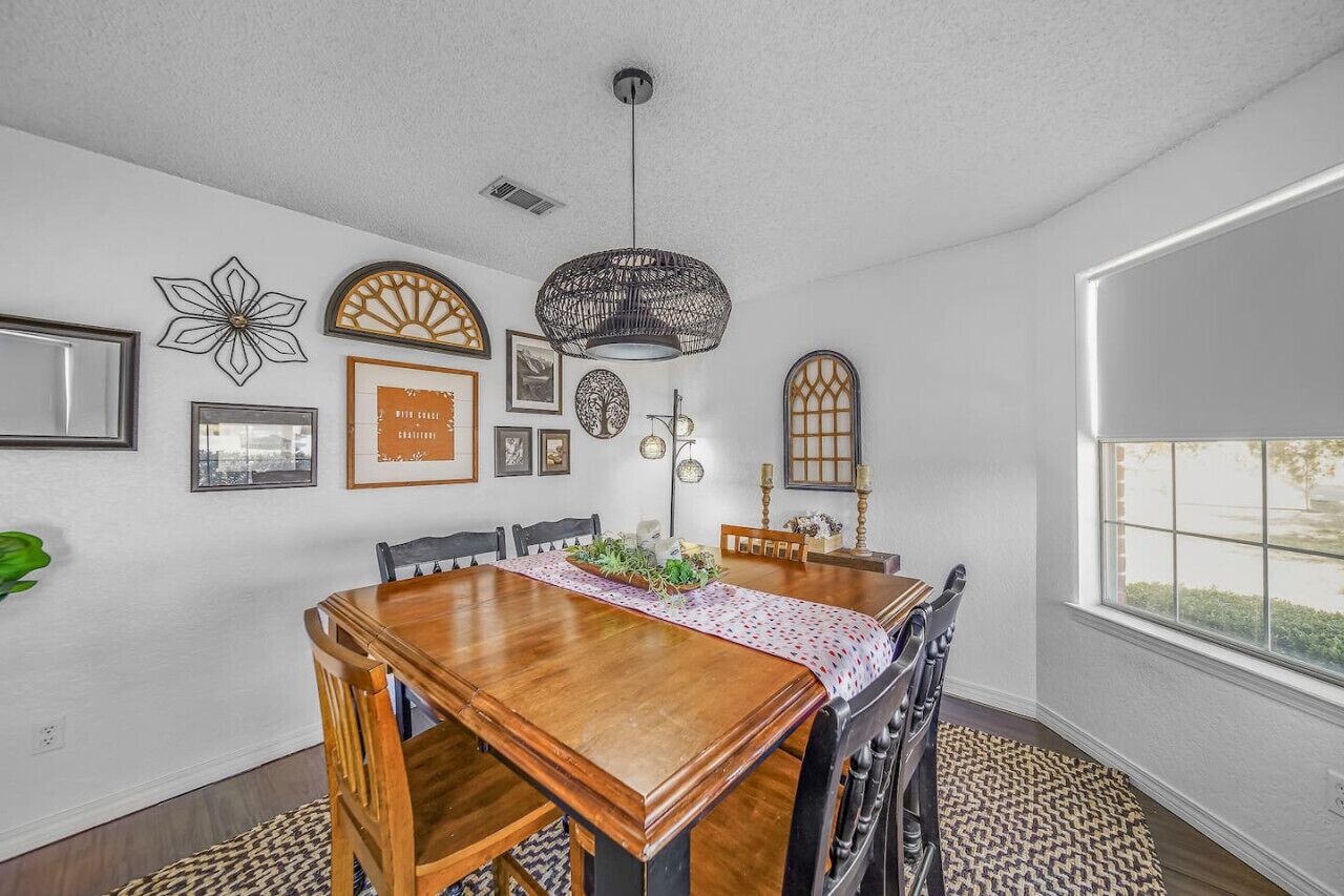 Un comedor con una mesa de madera, sillas y un camino de mesa estampado. Decoraciones en las paredes, incluyendo cuadros enmarcados y un diseño floral. Un ventanal grande proporciona luz natural.