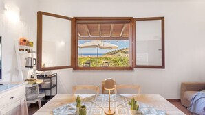 A room with a large window, a table with a patterned tablecloth, two chairs, and a view of a landscape with trees and a blue sky. - Casa Cactus Seaview with Pool (Tertenia)