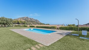 A rectangular swimming pool with a tiled deck, surrounded by well-maintained grass, a mountain in the background, and a street lamp. - Casa Cactus Seaview with Pool (Tertenia)