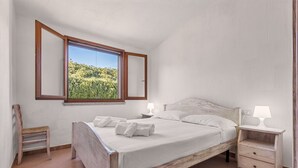 A bedroom with a wooden bed, two bedside tables with lamps, a chair, and a window with a view of greenery. - Casa Cactus Seaview with Pool (Tertenia)