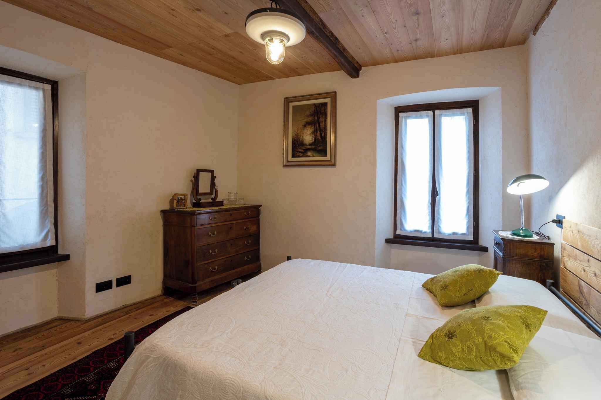 Een slaapkamer met een groot bed, twee ramen, een kaptafel, een schilderij en een nachtkastje met een lamp.