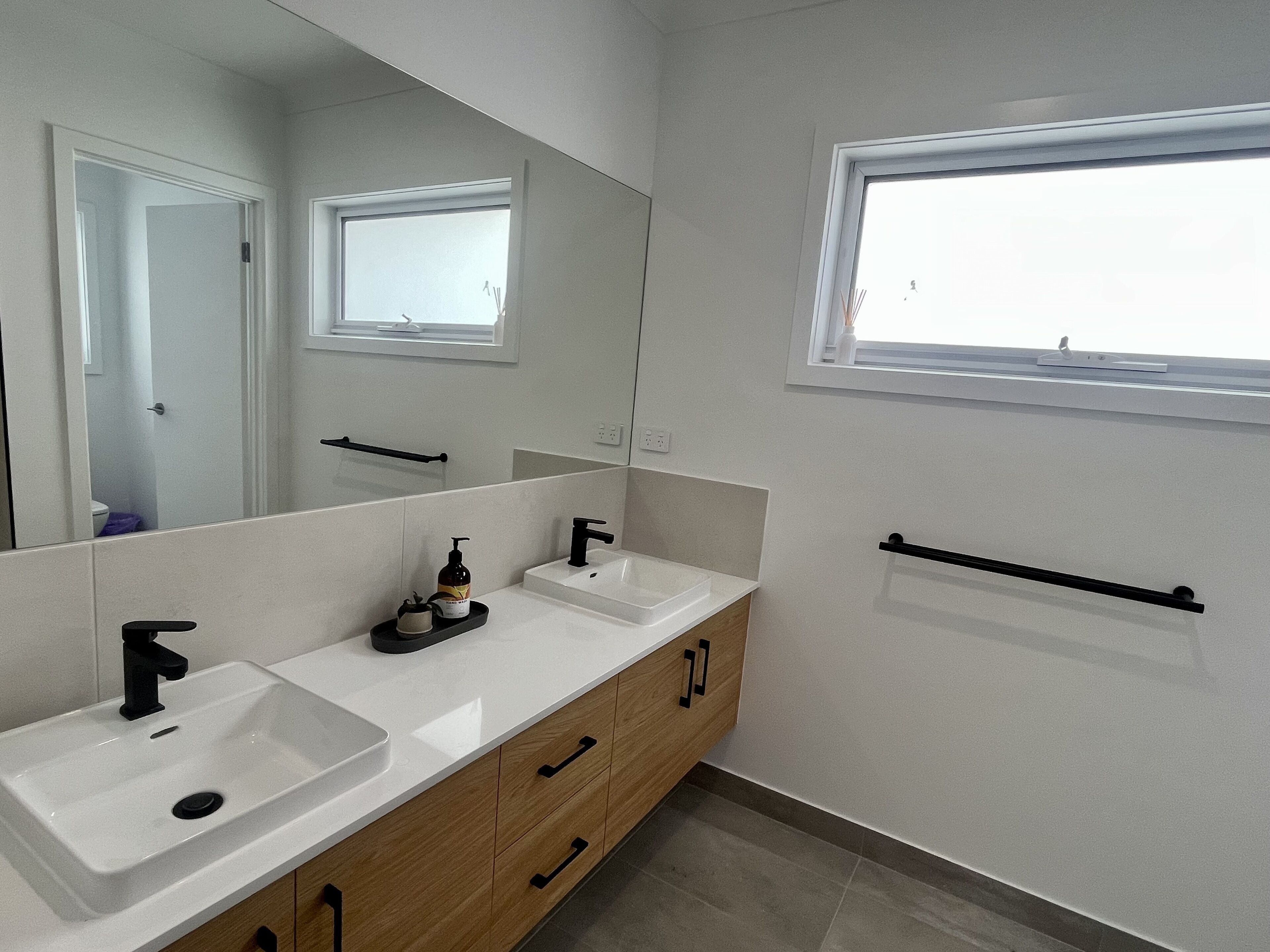 A modern bathroom with a large mirror, two sinks, wooden cabinets, and a window.
